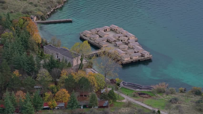 Aerial orbit over the Bay of Bones at Lake Ohrid after rain, capturing dramatic clouds, reflections on calm water and the historic prehistoric lakeside settlement.