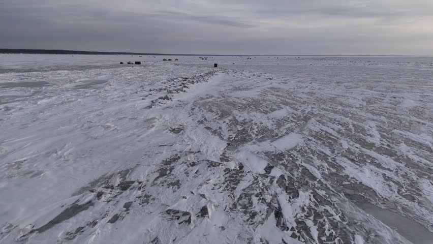 Drone flying forward over the frozen surface of Lake Winnipeg with partially snow covered ice. Ice fishing shacks appear in the distance across the winter lake landscape.