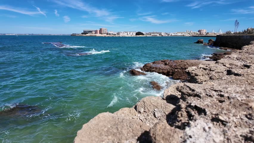 Waves roll and crash against a rugged rocky shoreline along the coast of Cádiz, Spain. Clear turquoise water and distant city skyline create a scenic Mediterranean seaside landscape.