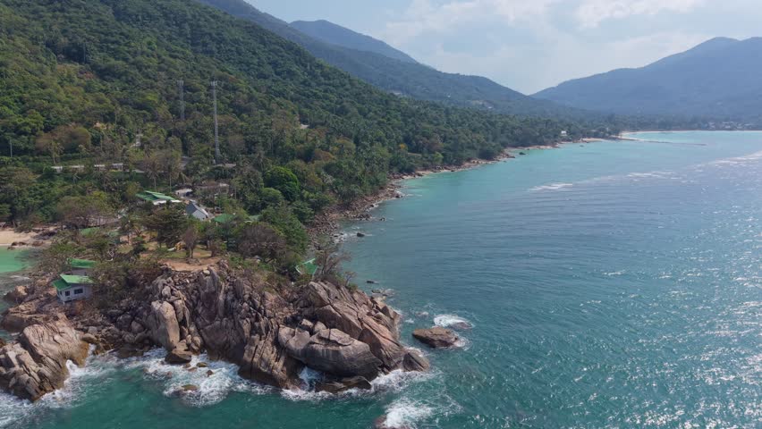 Drone push shot of a rocky coastline with tropical hills and clear ocean water in Thailand.
