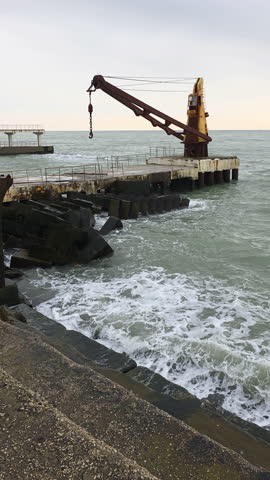 Old rusty harbor crane on a concrete pier with ocean waves crashing against the breakwater, creating a moody and industrial coastal seascape at dawn or dusk