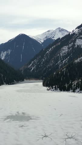 Majestic aerial view of the frozen kolsai lake during winter. The icy surface is surrounded by snow-dusted pine forests and mountains