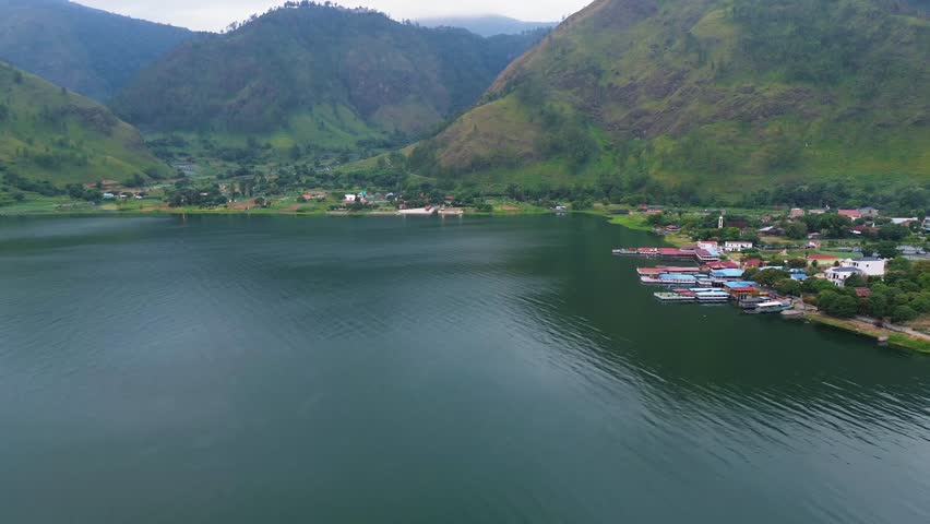 Aerial drone view of the vast Lake Toba landscape with surrounding hills, village houses, fish farms, and a small harbor visible from a distance as the drone slowly moves forward.