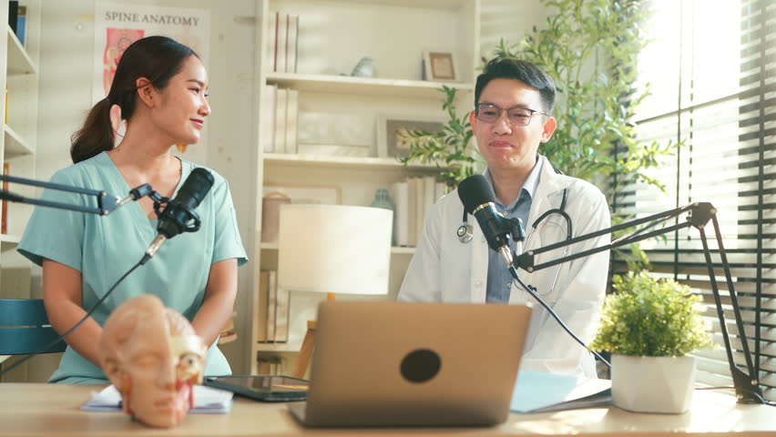 Asian medical doctor hosting a healthcare podcast interview with a nurse inside a bright clinic office. Professional consultation recording with microphones and laptop discussing treatment, patient