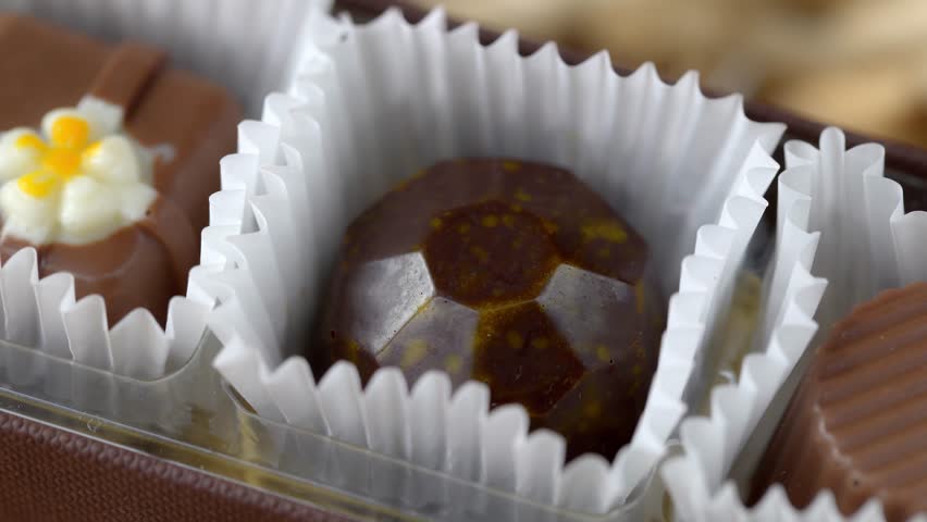 Macro close up of a hand selecting a chocolate candy from an assortment of pralines in a gift box. Sweet gourmet confectionery presented in paper cups, concept of indulgence, dessert choice