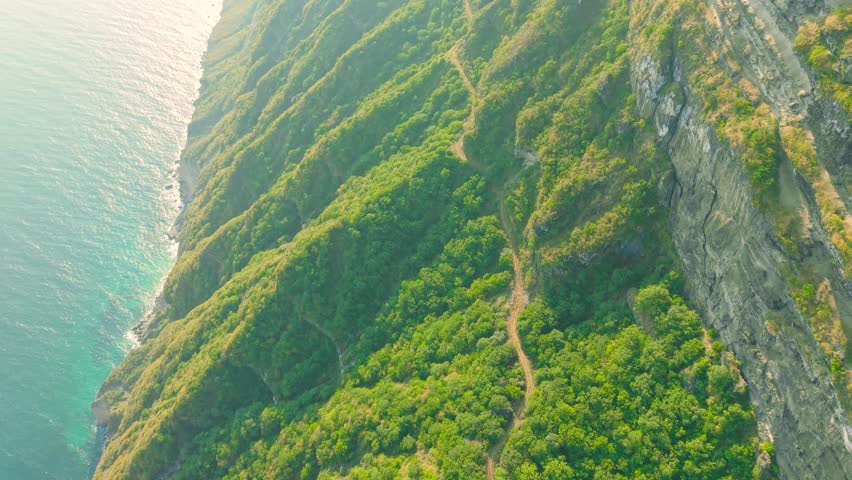 Aerial view of steep green cliffs and a narrow dirt road along the coast at Jabal Shaat on the border between Oman and Yemen.