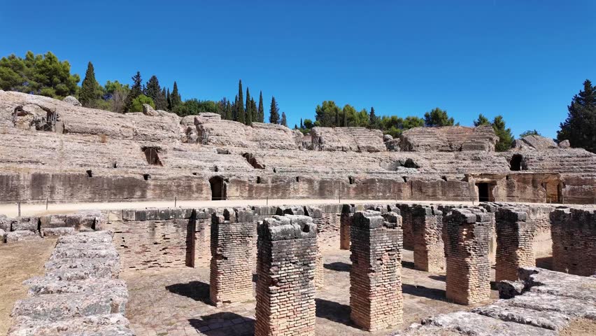 Wide view of the ancient Roman amphitheatre ruins at the Italica archaeological site near Seville, Spain. Historic stone seating and arena remain from the Roman Empire under clear blue skies.
