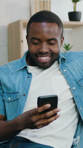 Handsome african man resting on comfortable sofa in beautifully decorated sitting room and is typing message on phone