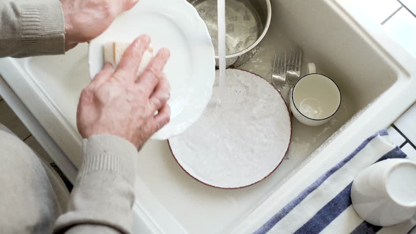 Man washes dishes in a kitchen sink with a sponge and liquid soap under running water. Contemporary domestic scene and modern masculinity. Concept of sharing household chores. Day light, close-up