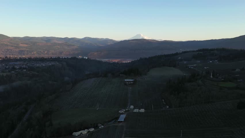 4K aerial drone footage flying over Hood River, Oregon during sunrise with Mount Adams visible in the distance and the Columbia River winding through the valley landscape. Soft morning light illuminates farmland, hills and rural terrain of the Pacific Northwest. Cinematic aerial b roll footage suitable for travel, tourism, nature documentaries, marketing videos, background visuals, presentations, media content and commercial projects showcasing scenic Oregon landscapes in the United States.