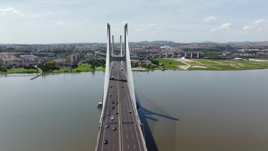 Aerial drone footage flying towards the majestic Vasco da Gama Bridge in Lisbon, Portugal. Cars and trucks cross the cablestayed bridge over the Tagus River on a bright, sunny day. Cityscape visible.