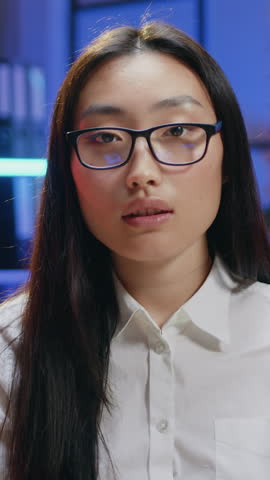 Asian woman office worker in glasses looking into camera with friendly face expression during overtime work at dark time in office