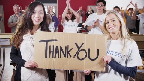 Two attractive female charity volunteers of Caucasian and Asian ethnicity hold up a "Thank You" sign and smile into the camera. Their fellow volunteers applaud and cheer in the background.Slow motion.