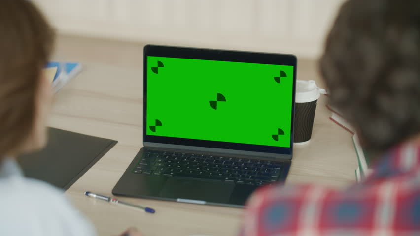 Over the shoulder view of two students collaborating on a project in a library or classroom, using a modern laptop with a chroma key green screen for custom content insertion