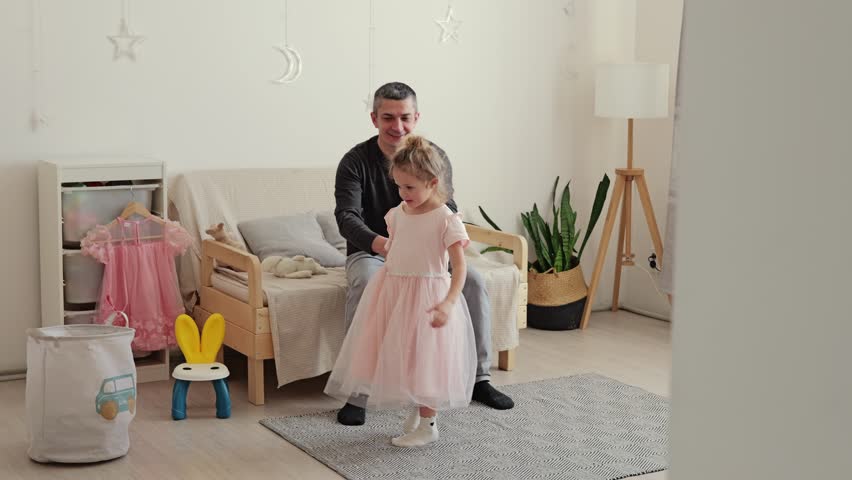A child dances happily while an adult watches and smiles in a simple room at home during the afternoon. Soft lighting creates a warm atmosphere