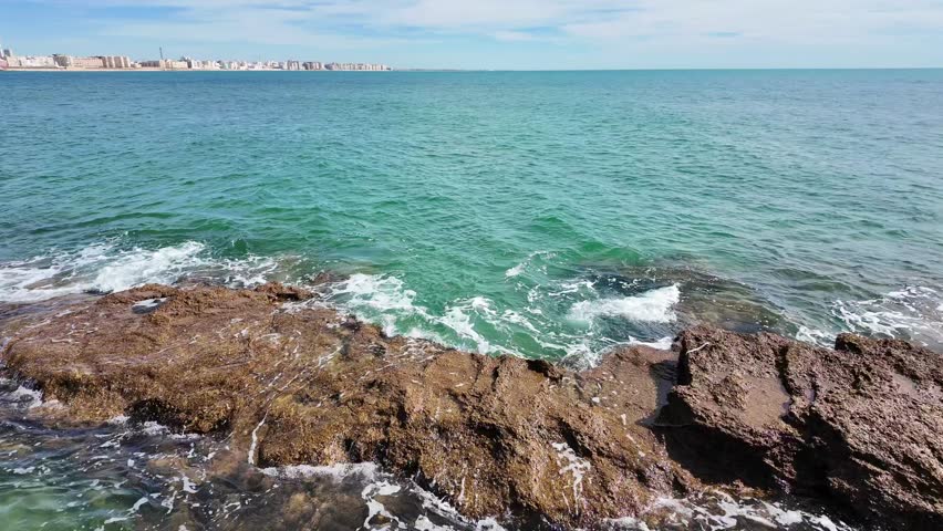 Waves roll and crash against a rugged rocky shoreline along the coast of Cádiz, Spain. Clear turquoise water and distant city skyline create a scenic Mediterranean seaside landscape.