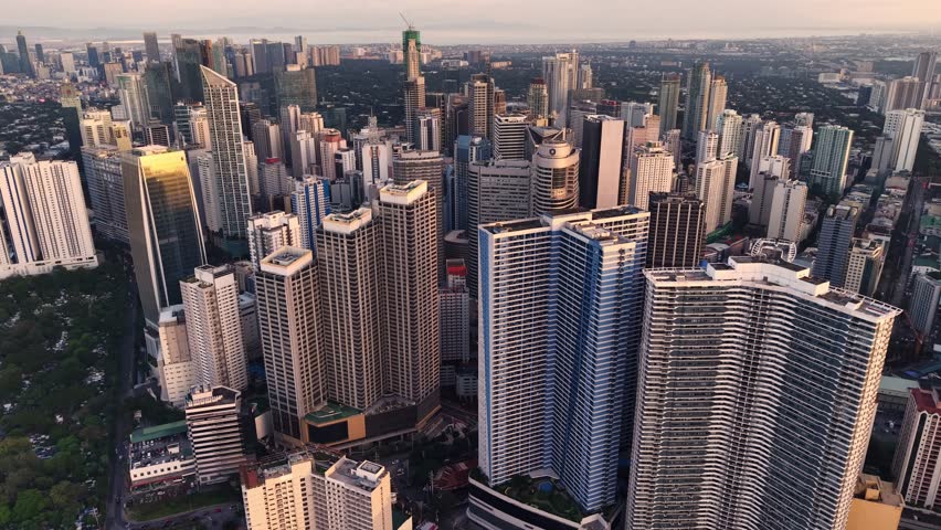 The drone ascends to high altitude, capturing an expansive panoramic view that encompasses a vast stretch of Metro Manila from dense low-rise residential neighborhoods in the foreground to distant tower clusters on the horizon, with Manila Bay visible along the left edge. The foreground is filled with a dense grid of low-rise rooftops in various colors and materials, stretching across the lower two-thirds of the frame and demonstrating the extraordinary urban density of Manila residential districts. Roads and narrow streets cut through the neighborhood fabric in visible linear patterns, creating a textured mosaic when viewed from this elevated vantage point. Two distinct high-rise clusters are visible at different distances, with a nearer group on the left and the taller Makati CBD towers further away on the center-right horizon. Manila Bay appears as a pale reflective expanse along the left portion, where cargo ships are visible as small dark shapes against the luminous water surface. The sunset creates a warm gradient across the horizon, blending golden and peach tones that illuminate wispy cirrus clouds in the upper sky, while lower portions of the city fall into cooler blue-gray tones of approaching dusk.