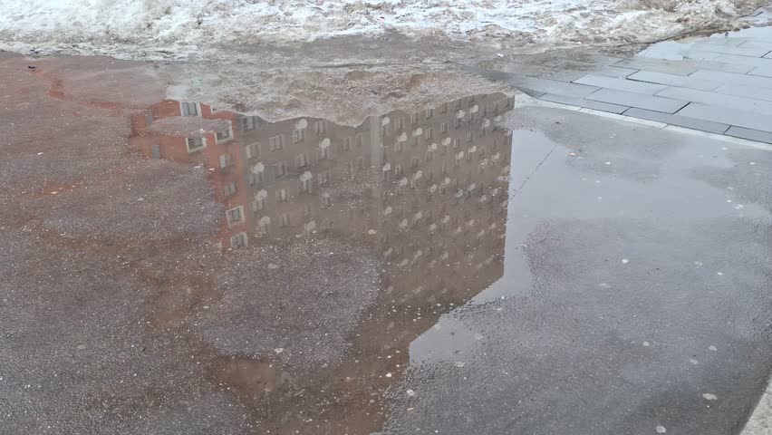 Pedestrians walk past large puddles and melting snow on city sidewalk. Wet asphalt reflects residential building. People feet move through slushy street, seasonal transition, urban weather conditions