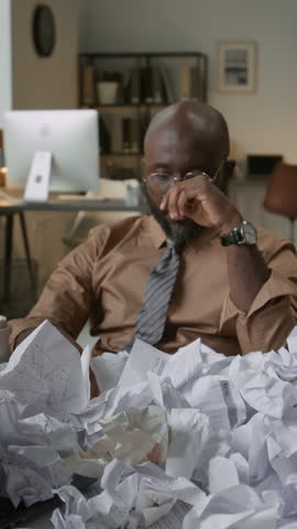 Vertical shot of frustrated African American man removing glasses and rubbing eyes while sitting at messy desk covered with crumpled papers in office