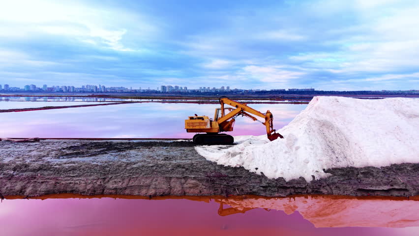 Side view of a yellow hydraulic excavator on a salt ridge with city buildings in the background and pink water reflections. Industrial excavator processing sea salt at evaporation site.