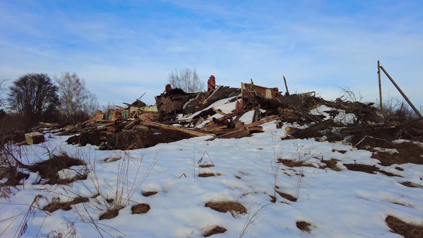 A building lies in ruins after a heavy snowstorm. Wooden planks and debris are scattered across the snowy ground. The sky is clear, and trees are visible in the background.
