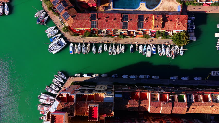 Top down aerial shot of white yachts and boats docked along a pier with red tiled roofs. Boats moored in marina with turquoise water.