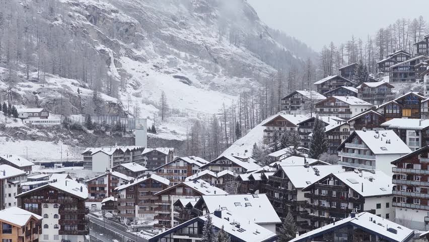 Aerial pan of Zermatt in the Swiss Alps with chalets, apartments, and a church steeple, illustrating alpine scenery, tourism, and mountain village life.