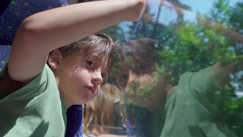 Curious young boy rides a bus, leaning on window and gazing outside, thoughtful expression reflected in glass, seated on patterned seat, forest landscape in background