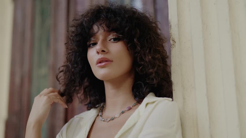 Slow motion portrait of a beautiful young woman with curly hair and elegant makeup posing sensually, gently touching her neck and looking at the camera with a captivating and alluring gaze