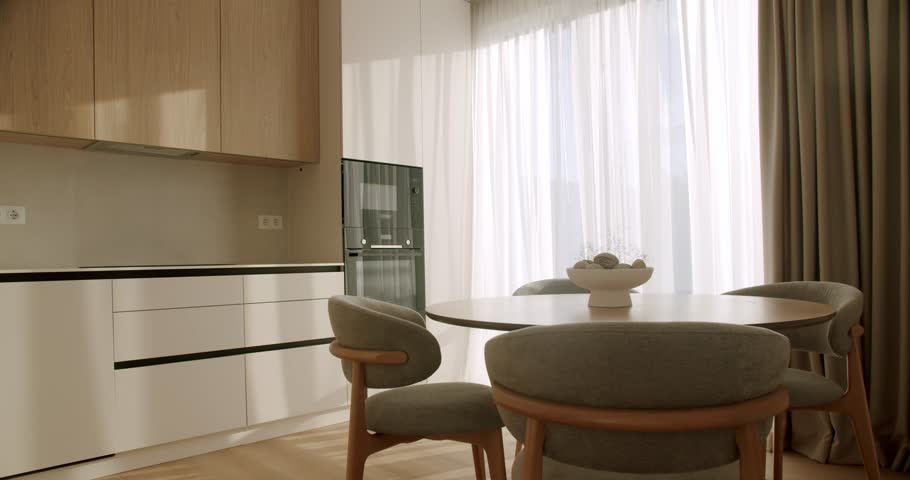 Bright and spacious kitchen interior with minimalist wooden cabinets and a contemporary dining set next to a large window with sheer curtains, bathed in beautiful morning sunlight