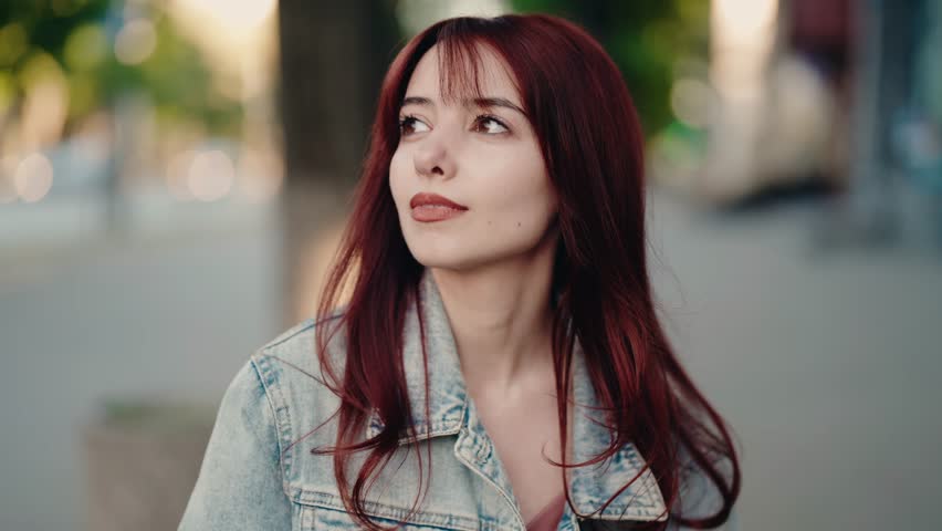 Charming young redhead woman with a stylish haircut wearing a denim jacket walking outdoors in the city street with a thoughtful expression, enjoying a moment of quiet contemplation