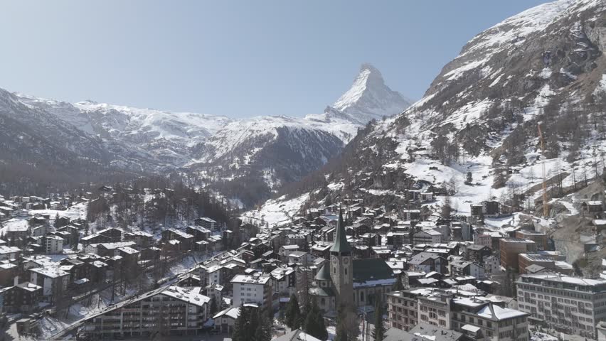 Cinematic drone footage of the iconic Matterhorn mountain rising above the picturesque alpine town of Zermatt, Switzerland. The famous church in the village foreground sits beneath the dramatic snow covered peak, creating a classic Swiss Alps landscape. Raw aerial footage capturing a peaceful winter atmosphere with pristine snow, charming chalets, and the legendary pyramid shaped Matterhorn often associated with the Toblerone mountain. Perfect for travel, holiday, winter, romantic, and cinematic storytelling content.