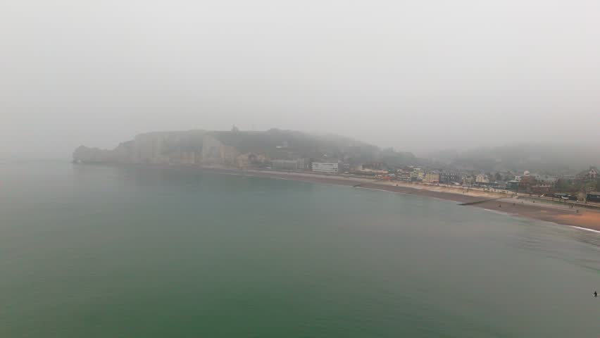 Aerial slow pan tracking tilt of coastal cliffs and beach revealing small town on foggy overcast shoreline.