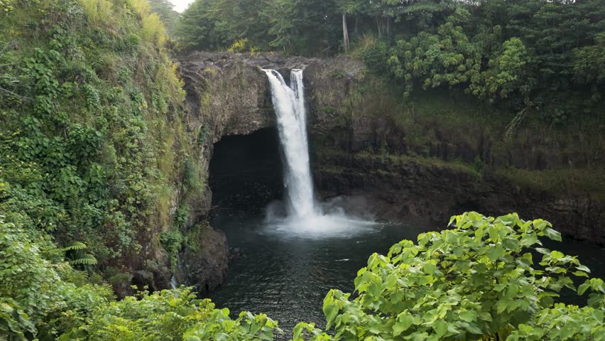 A stunning wide shot of the famous Rainbow Falls surrounded by dense tropical rainforest and vibrant green vegetation, Hawaii Island. High quality 4k footage