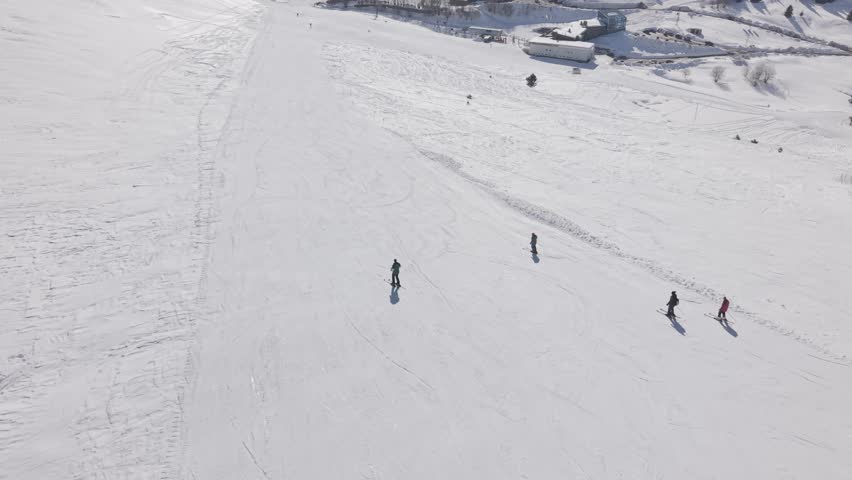 Aerial tracking shot following people skiing down a wide snow-covered slope at a winter mountain resort