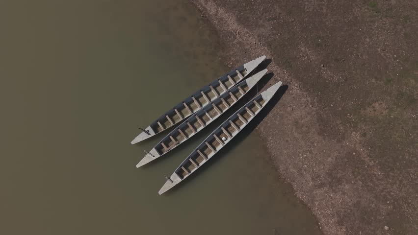 Aerial top-down forward tracking shot of three empty traditional rowboats moored on a muddy lake shore