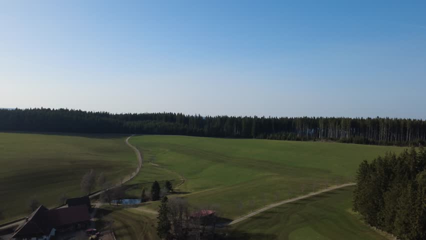 Smooth aerial panorama of peaceful rural farmland landscape on a bright sunny day. Scenic countryside with fields, forest edges, small roads and scattered houses viewed from above. Calm natural environment with wide open agricultural land and gentle rolling terrain.
