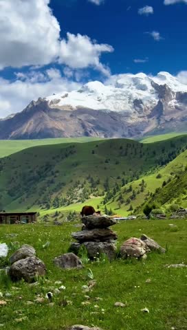 Wide mountain landscape with green grass meadow and snow covered peaks under cloudy sky.
