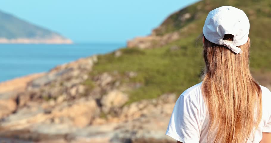 Back view of woman in white cap looking at scenic ocean landscape. Rocky coastline and blue water under bright sun. Concept of travel, freedom, and nature exploration.