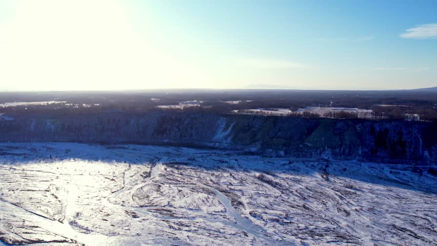 Medium altitude drone flight directly into winter sun over frozen braided Matanuska River flats near Palmer, AK. Dramatic shadowed bluff dominates right, sun glare illuminates vast icy channels below.