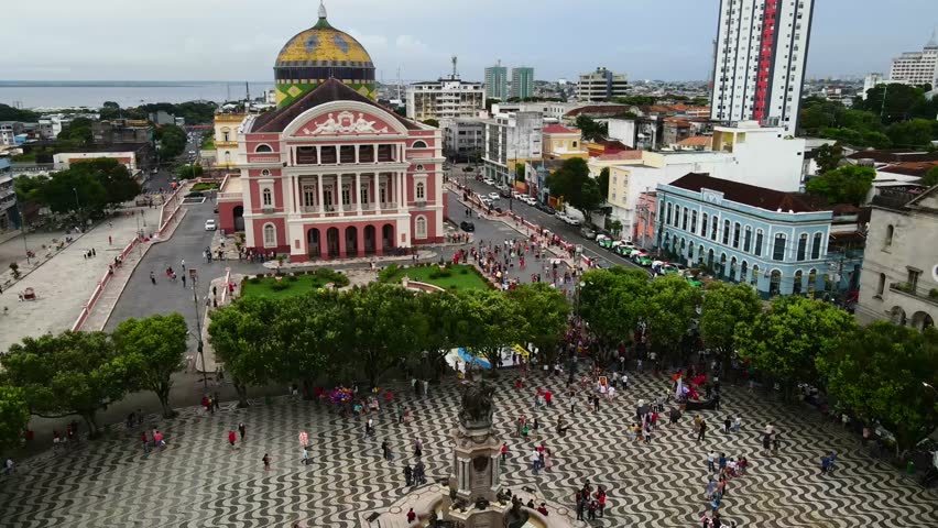 Aerial orbit of Teatro Amazonas and Largo de São Sebastião square in Brazil. Crowds of people are gathered around the central fountain during cloudy afternoon.