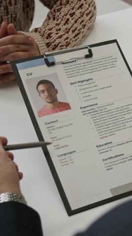 Vertical shot of unknown recruiter looking through resume of positive young man and speaking with him while conducting job interview in office