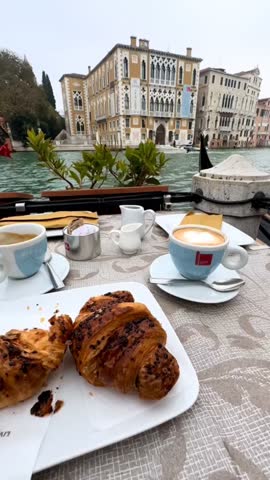 Cinematic 4K vertical footage capturing a traditional Italian breakfast on a charming terrace overlooking the famous Grand Canal in Venice. A cup of fresh coffee and flaky croissants are served on a café table while iconic gondolas glide along the canal beside historic Venetian architecture. This elegant morning scene highlights the authentic atmosphere of a Venetian café lifestyle, perfect for travel marketing, tourism promotion, food and beverage advertising, luxury lifestyle content, and European city travel visuals.