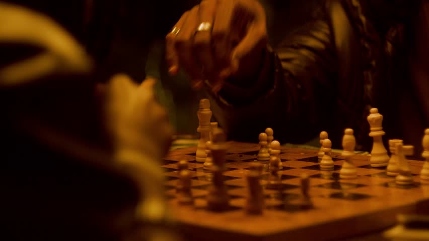 Detailed close-up of men's hands moving chess piece during an intense night game. Concepts of strategy, calculated risk, decision making, and urban competition with warm cinematic lighting.