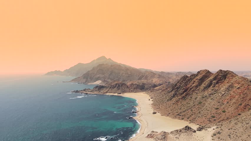 Aerial view of the coastline featuring rugged mountains, sandy beach, and turquoise ocean water under a hazy orange sky, Socotra, Yemen.