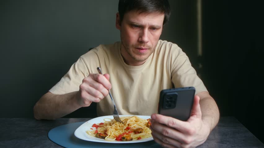 Serious man with phone addiction eating italian pasta for lunch, browsing social media, watching videos reading news feeds, completely absorbed by screen. Addicted man eating spaghetti scrolling phone