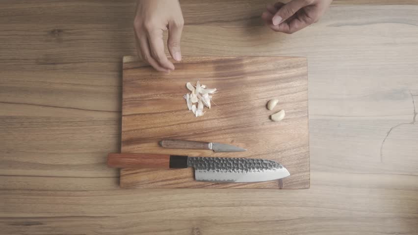 Close-up view of food preparation showing peeling, slicing, and mincing fresh garlic cloves on a cutting board in a kitchen. Demonstrates essential culinary techniques used in home cooking and professional kitchens. Ideal for illustrating ingredient preparation, healthy cooking, meal prep, seasoning preparation, and kitchen workflow for recipes and cooking tutorials.