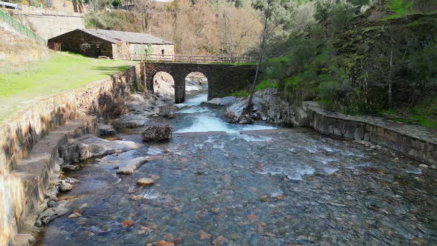 Drone shot of ancient stone bridge over clear river with rocky riverbed and traditional house in Extremadura Spain. Scenic European travel destination and heritage landscape.