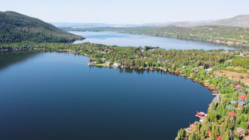 High aerial view of Grand Lake Colorado featuring waterfront houses, wooded peninsulas, deep blue water, and layered mountain scenery in a peaceful summer resort setting