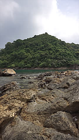 Rocky shoreline with flowing water in the foreground and a lush green hill under a cloudy sky